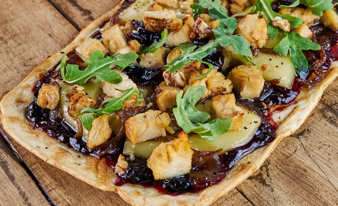A flatbread topped with diced chicken, onions, cheese, arugula and pecans sitting on a wood table