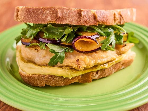 Grilled chicken on sourdough with pistachio butter, marinated plums, and arugula on a green plate, on a wooden surface