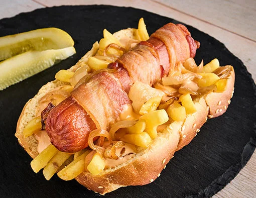 A bacon-wrapped hot dog topped with fries and grilled onions, served with pickles on a black plate