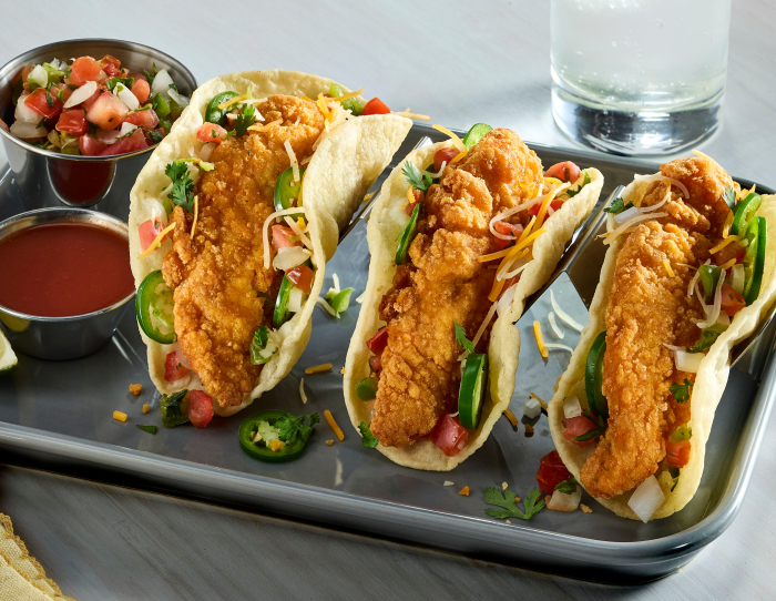 Three gluten-free breaded chicken tender tacos with lettuce, jalapeños, pico de gallo, and dipping sauces on a metal tray