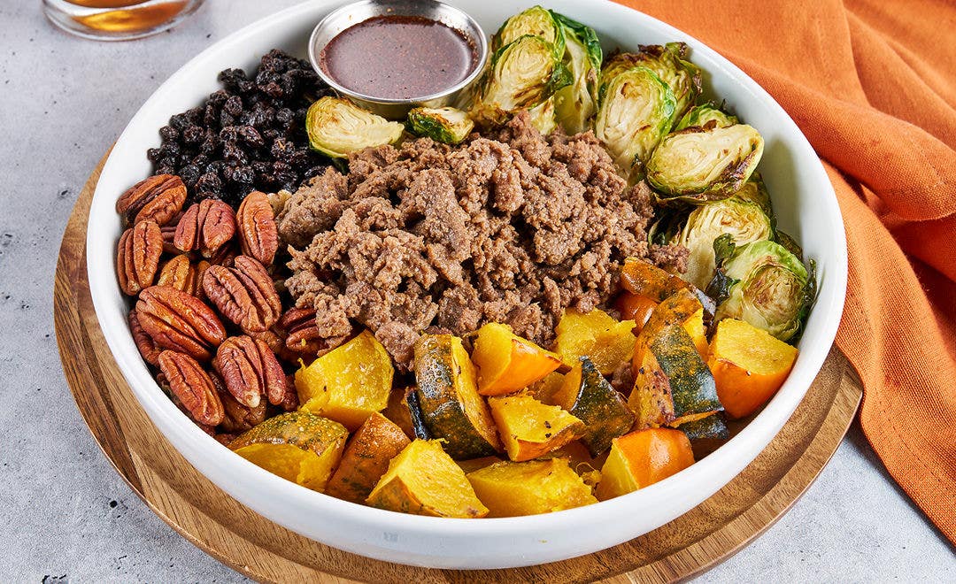 A bowl loaded with sliced beef, acorn squash, pecans, black currants, Brussels sprouts and sauce in a metal ramekin