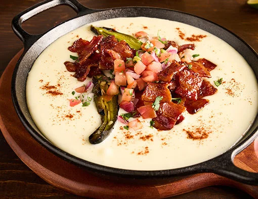 Skillet of creamy queso topped with bacon, pico de gallo, jalapeño, and chicharrones on the side