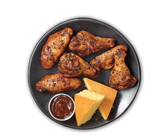 Overhead view of a black plate with six seasoned chicken wings, a small cup of barbecue sauce, and two triangular cornbread slices