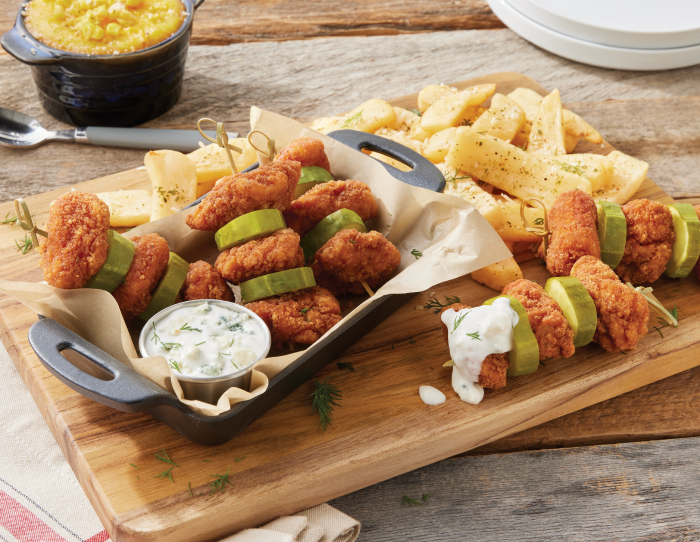Skewers of spicy breaded chicken bites layered with pickle slices, served with seasoned fries and a creamy dipping sauce on a wooden board