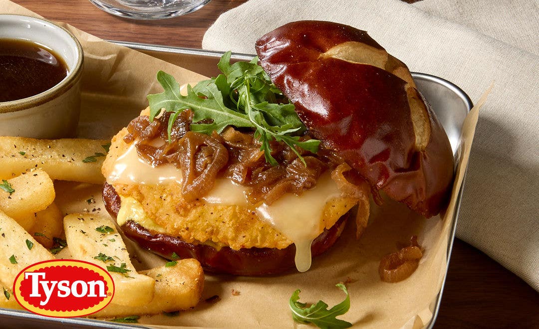 A metal tray with a breaded chicken breast on an open pretzel bun smothered in melted cheese and caramelized onions 