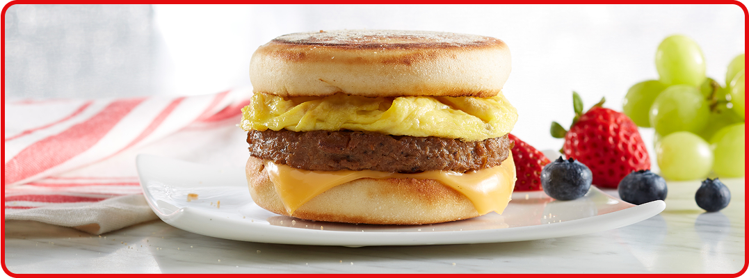 A sausage, egg and cheese breakfast sandwich on an English muffin. Sides of blueberries, strawberries and grapes sit to the side of the plate. 