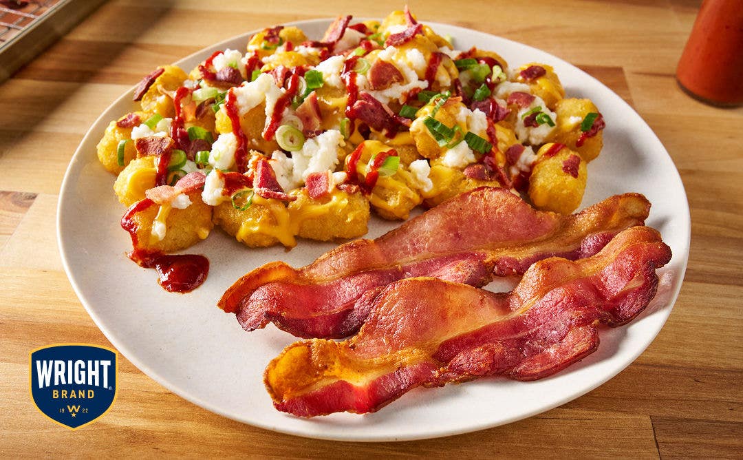 A plate with potato tots covered in cheese, bacon pieces, scrambled eggs, scallions and hot sauce with a side of bacon slices 