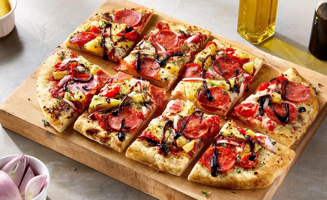 Pepperoni flatbread pizza with cherry tomatoes, pineapple, cheese and a balsamic glaze on a cutting board