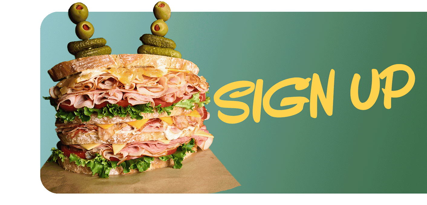 A fully loaded dagwood sandwich on parchment paper stacked high with a variety of sliced meats, cheeses, lettuce and tomato held together with olive and pickle spears  