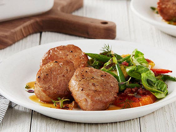 Orange and Thyme Pork Medallions With a Side of Greens on a White Plate and Wooden Table