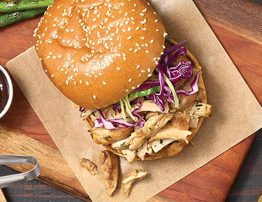Close-up of a smoked chicken sandwich on a sesame bun with sliced chicken, purple cabbage slaw, and sauce on parchment over a wooden board