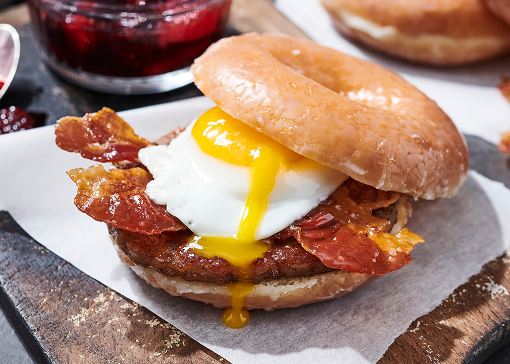 Breakfast sandwich with sausage, bacon, and a fried egg on a glazed donut, served on a wooden board