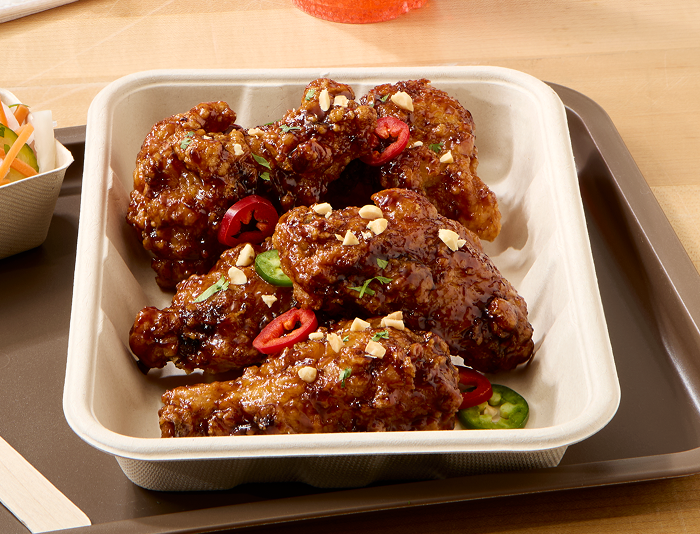 Fully cooked glazed chicken wings in a takeout tray, garnished with sliced red and green chili peppers and chopped peanuts on a serving tray
