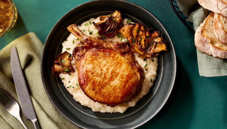 Golden-brown, bone-in pork chop over creamy risotto with roasted mushrooms in a dark bowl