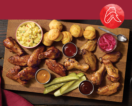 Platter of smoked chicken wings with dipping sauces, pickles, cornbread bites, potato salad, and pickled onions on a rustic wooden board