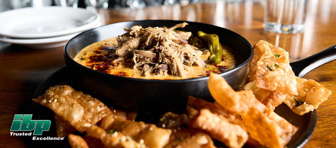 A bowl of queso dip topped with pulled pork and shishito peppers on a plate with wonton chips 