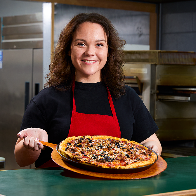 A smiling woman in a red apron holds a deep-dish pizza on a wooden peel in a restaurant kitchen