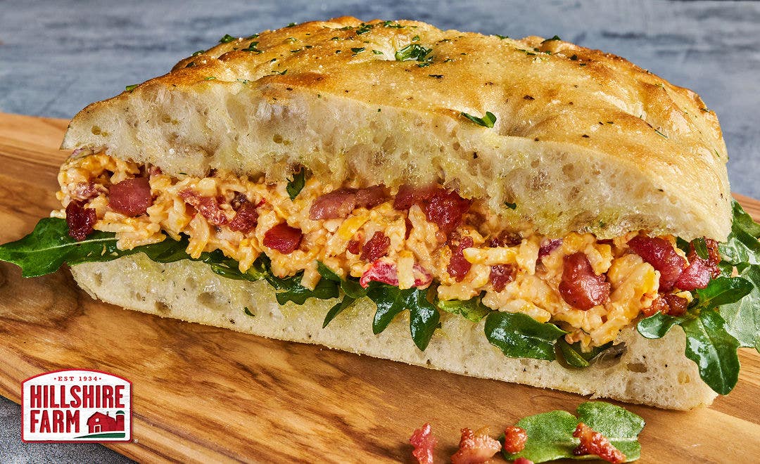 A cutting board holding a bacon, arugula and pimento cheese sandwich on focaccia bread 
