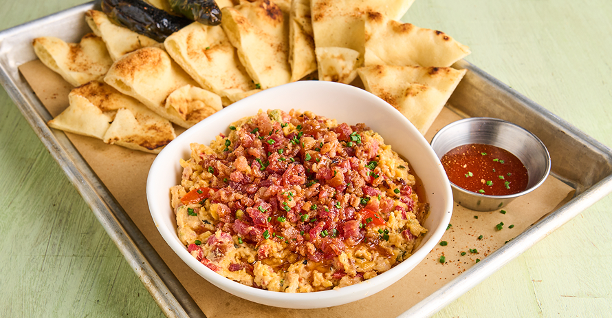 A bowl of dip topped with bacon, served with pita bread and peppers on a tray