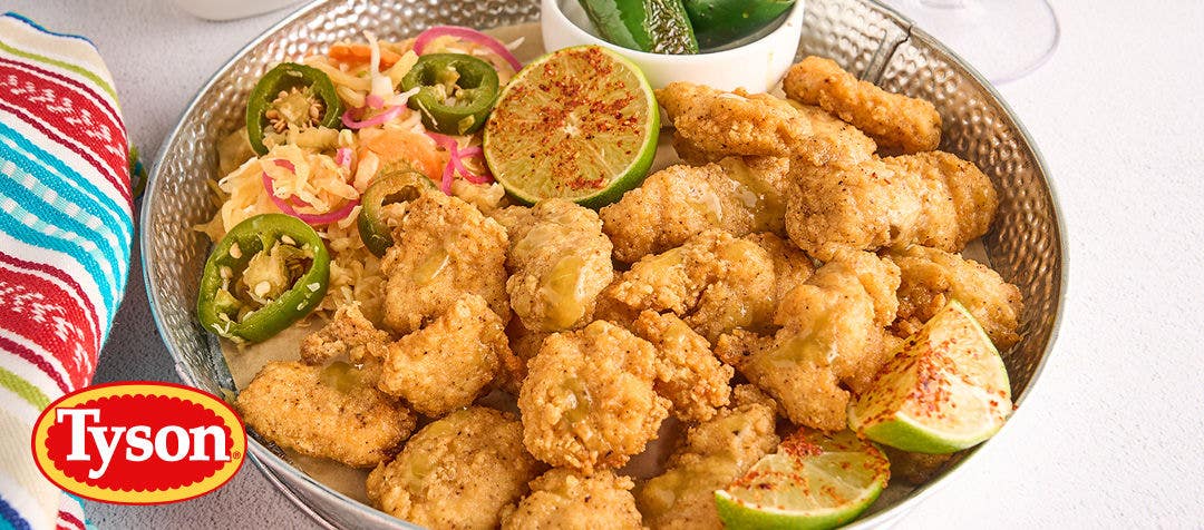 A metal bowl filled with crispy, breaded chicken chunks, sliced jalapeños, lime wedges and slaw       