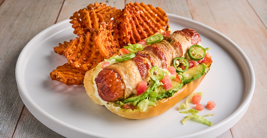 A bacon-wrapped hot dog topped with avocado, lettuce, tomatoes, and jalapeños, served with sweet potato fries