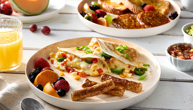 Breakfast quesadilla with a side of sausage links and fruit on a white plate