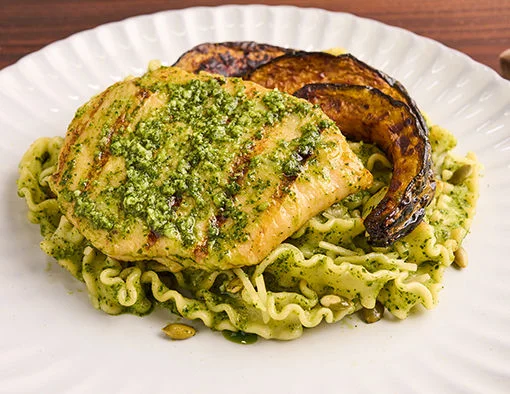 Grilled chicken on mafaldine pasta with pepita pesto, roasted squash, and arugula on a white plate, on a rustic wooden surface