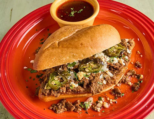 Birra cheesesteak with meat, cheese, and jalapeños on a red plate with a side of dipping sauce