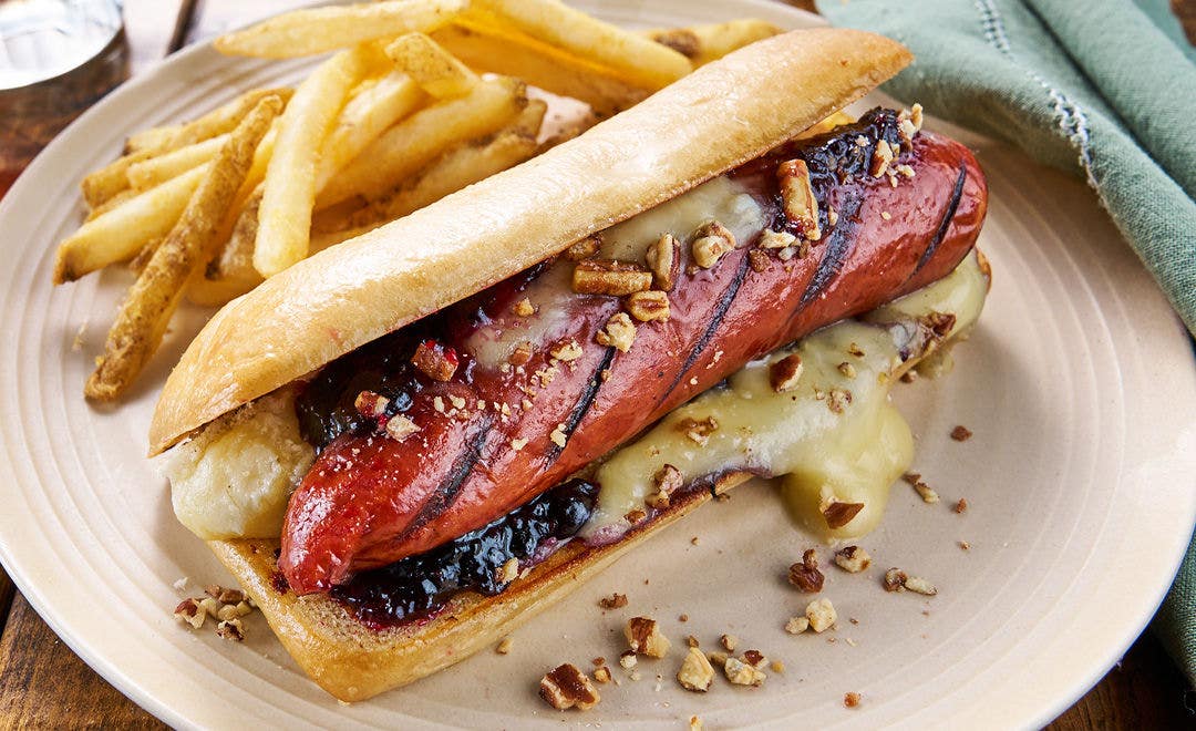 A plate with French fries plus a sausage on a roll with melted cheese, fruit chutney and a sprinkling of chopped walnuts 