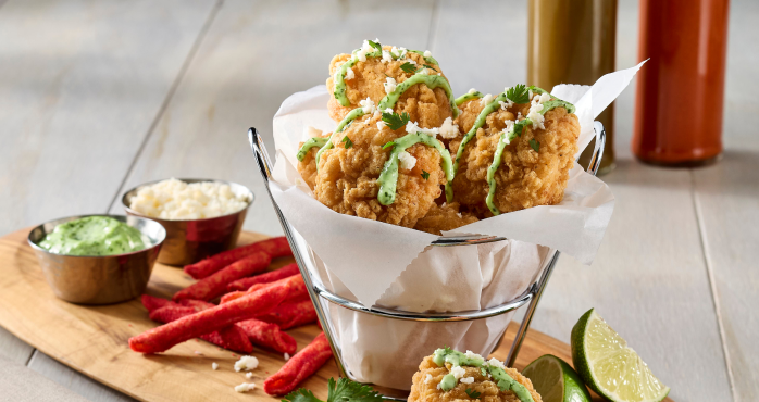 A basket of fried chicken bites with green sauce, cheese, and red snacks