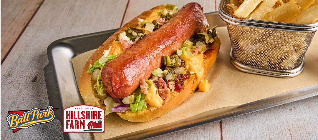 A metal tray with a hot dog loaded with chili, cheese, slaw, bacon and relish along with a side of fries 