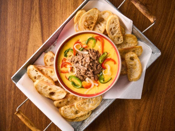 A bowl of creamy cheese dip topped with steak and peppers, served with toasted bread slices on a tray