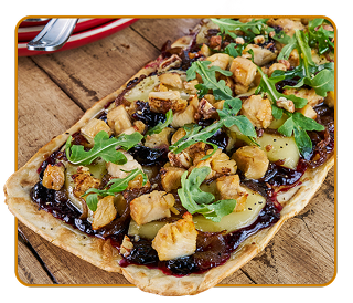 A flatbread topped with diced chicken, onions, cheese, arugula and pecans sitting on a wood table 