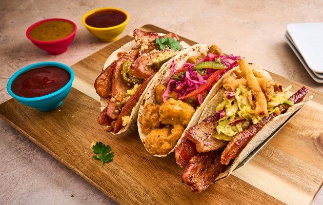 Three assorted tacos on a wooden board with meat, slaw, and colorful dipping sauces