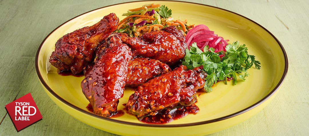 A yellow plate on a table with red sauced chicken wings, slaw, pickled onions and fresh cilantro    