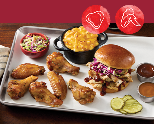 Tray of smoked chicken wings with a pulled chicken sandwich, mac and cheese, coleslaw, pickles, and barbecue sauces on a metal platter