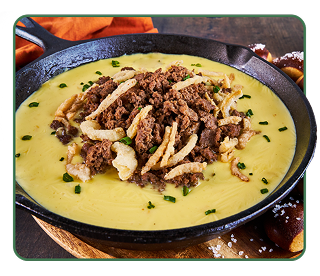 A cheese fondue in a black skillet layered with sliced beef, roasted garlic, fried shallots and chives 