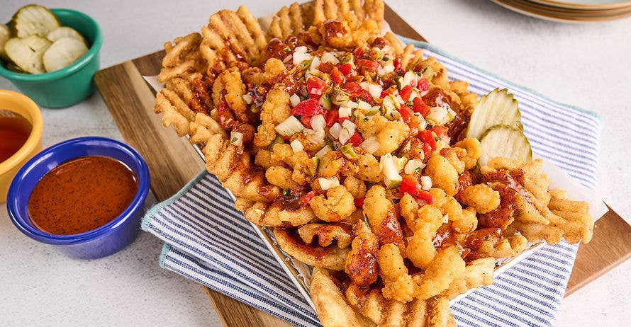 Waffle fries topped with crispy chicken bites, red sauce, relish, and pickles in a basket