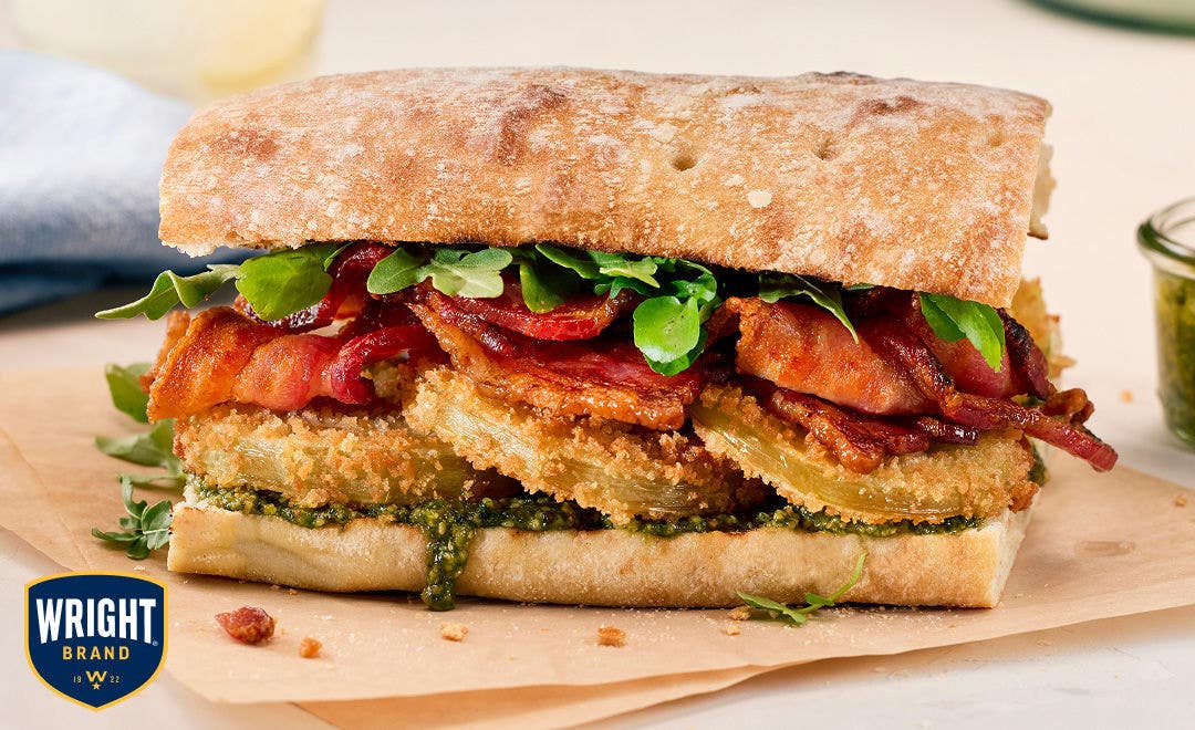 A whole wheat roll on parchment paper stuffed with sliced bacon, fried green tomatoes, arugula and pesto    