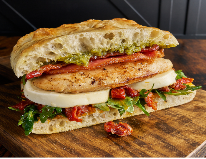A grilled chicken breast sandwich layered with thick mozzarella cheese, sliced bacon, pesto and arugula resting o a wooden table   