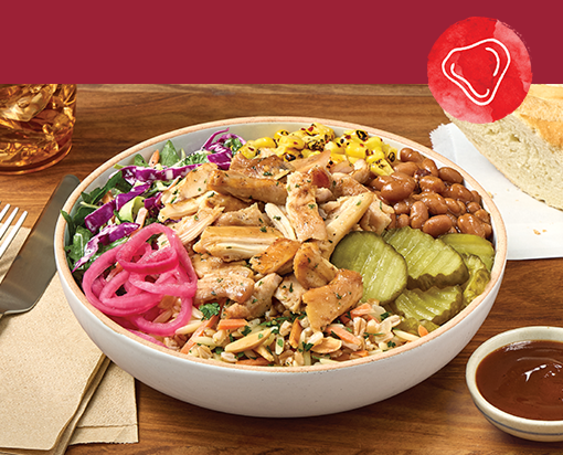 Bowl of smoked chicken grain bowl with sliced chicken, beans, pickles, pickled onions, slaw, grains, and barbecue sauce on wooden table