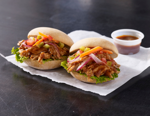 Two small sandwiches of pulled barbecue chicken on slider buns with red onions, carrots, relish and lettuce on a napkin 