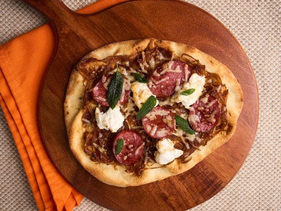 Flatbread with sausage, caramelized onions, ricotta, shredded cheese, and sage on a wooden board