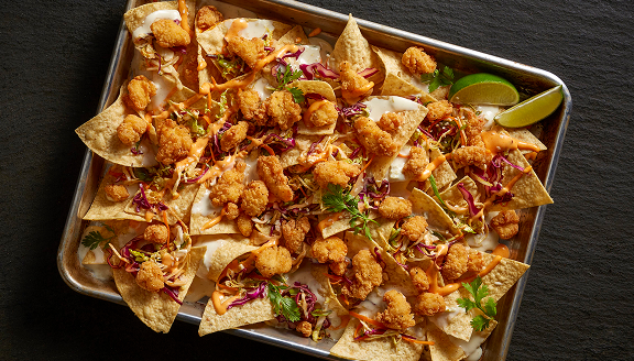 A sheet pan of loaded nachos with mini chicken bites, slaw, and sauces