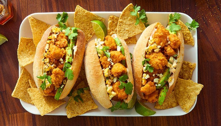 Three crispy chicken sandwiches with avocado, corn salsa, and cilantro on a platter with tortilla chips