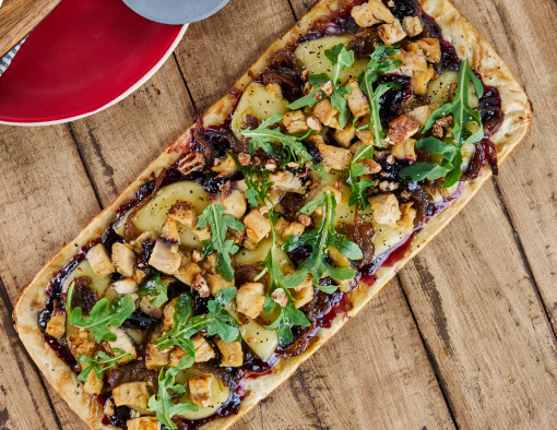 A flatbread topped with diced chicken, onions, cheese, arugula and pecans on a wood table