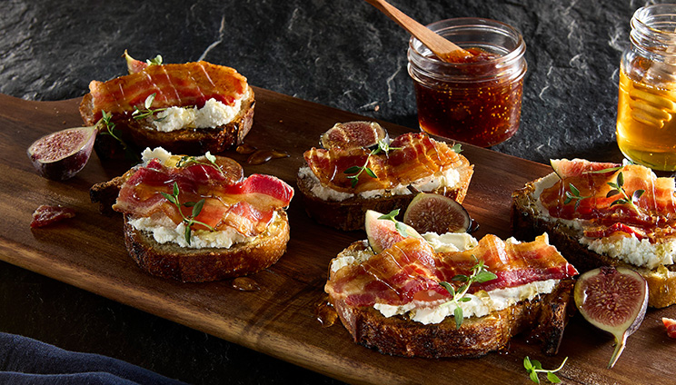 Crostini with goat cheese, bacon, figs, and honey on a wooden board, garnished with thyme, with jars of fig jam and honey in the background