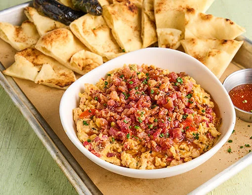 A bowl of dip topped with bacon, served with pita bread and peppers on a tray