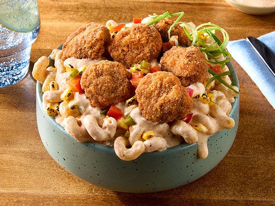 A bowl of mac & cheese topped with pickles, tomato, spicy breaded beef bites, and drizzled with a spicy buffalo ranch.