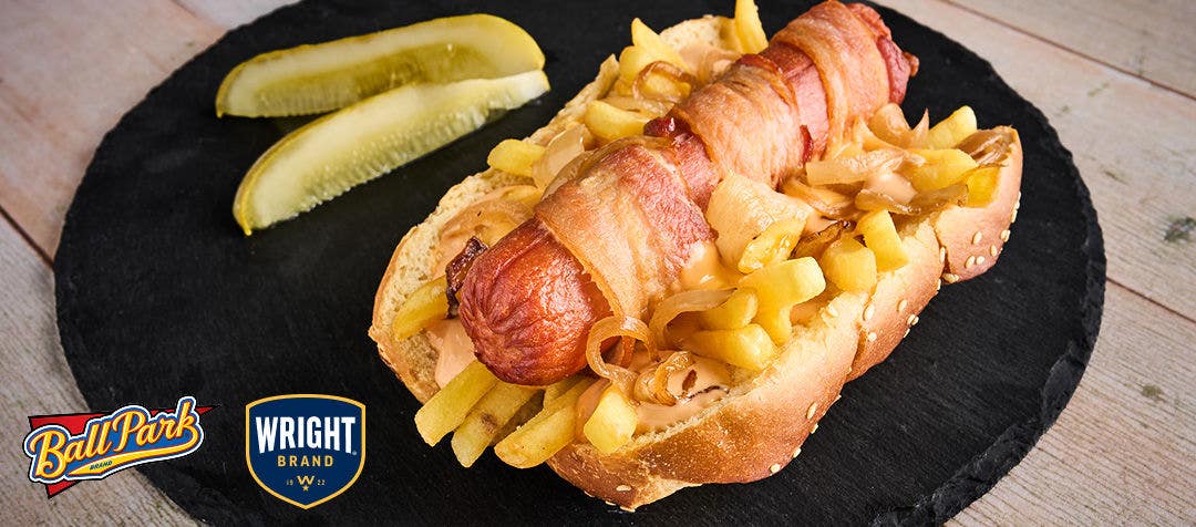 A black plate with a bacon-wrapped hot dog on a bun with French fries and grilled onions and pickle spears on the side   